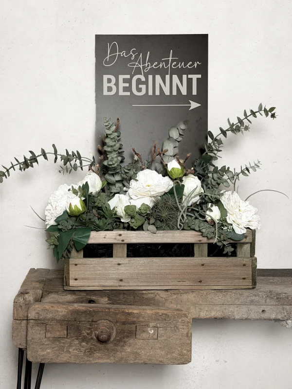 Vintage Holzkiste mit Textilblumengesteck und Schild "Das Abenteuer beginnt"